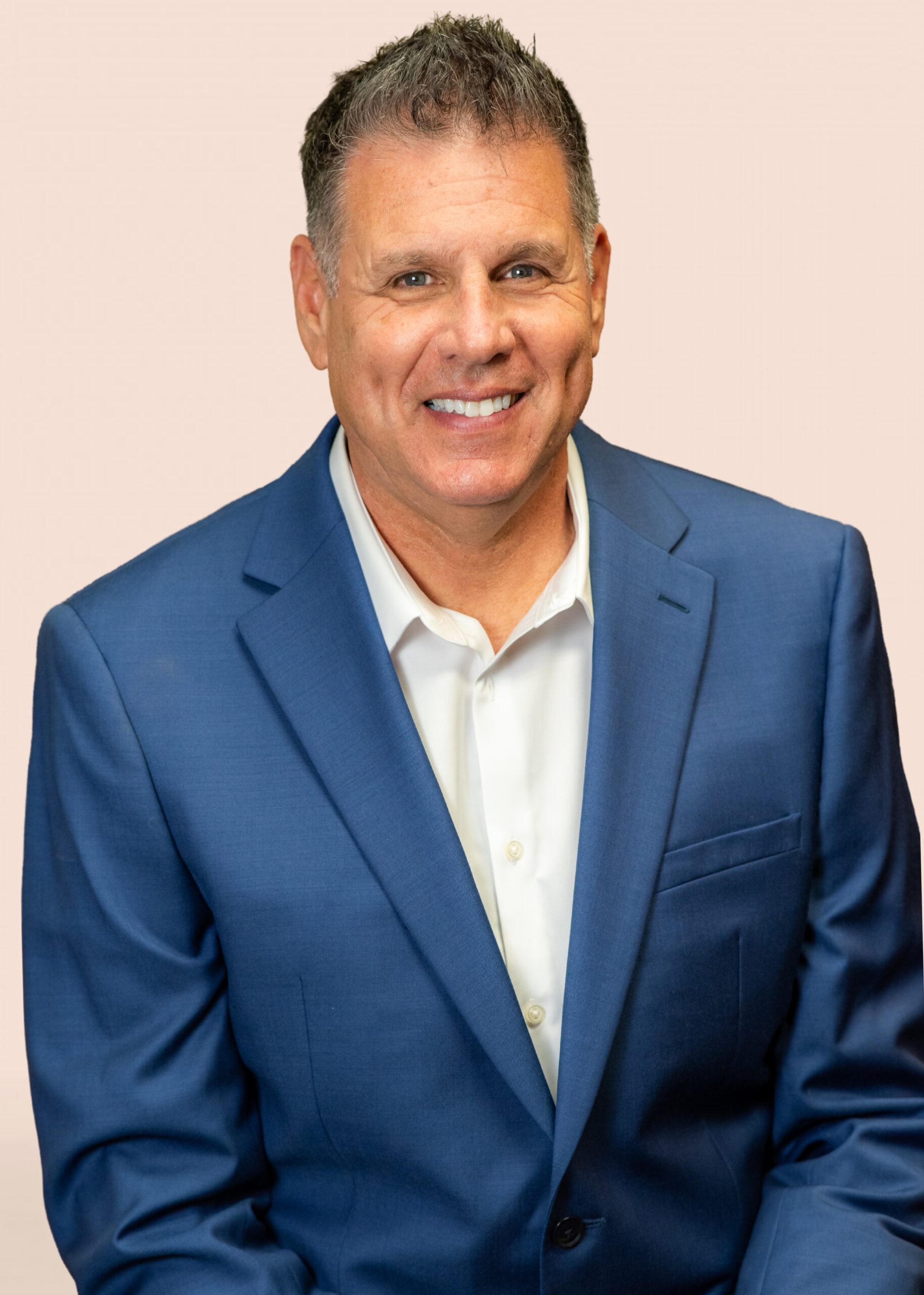 Dr. Jose with short dark hair smiles at the camera. He is wearing a blue suit jacket over a white collared shirt and is seated in front of a plain, light-colored background.