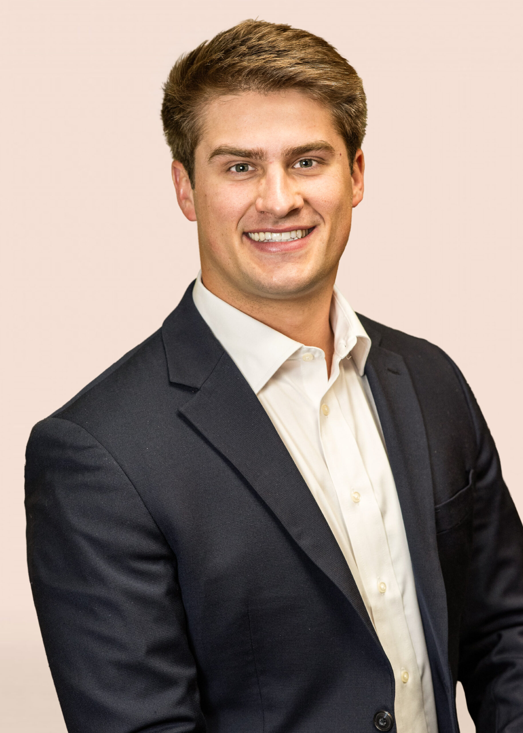 Dr. Taylor with short brown hair wearing a dark suit jacket and white dress shirt smiles in front of a plain, light-colored background.