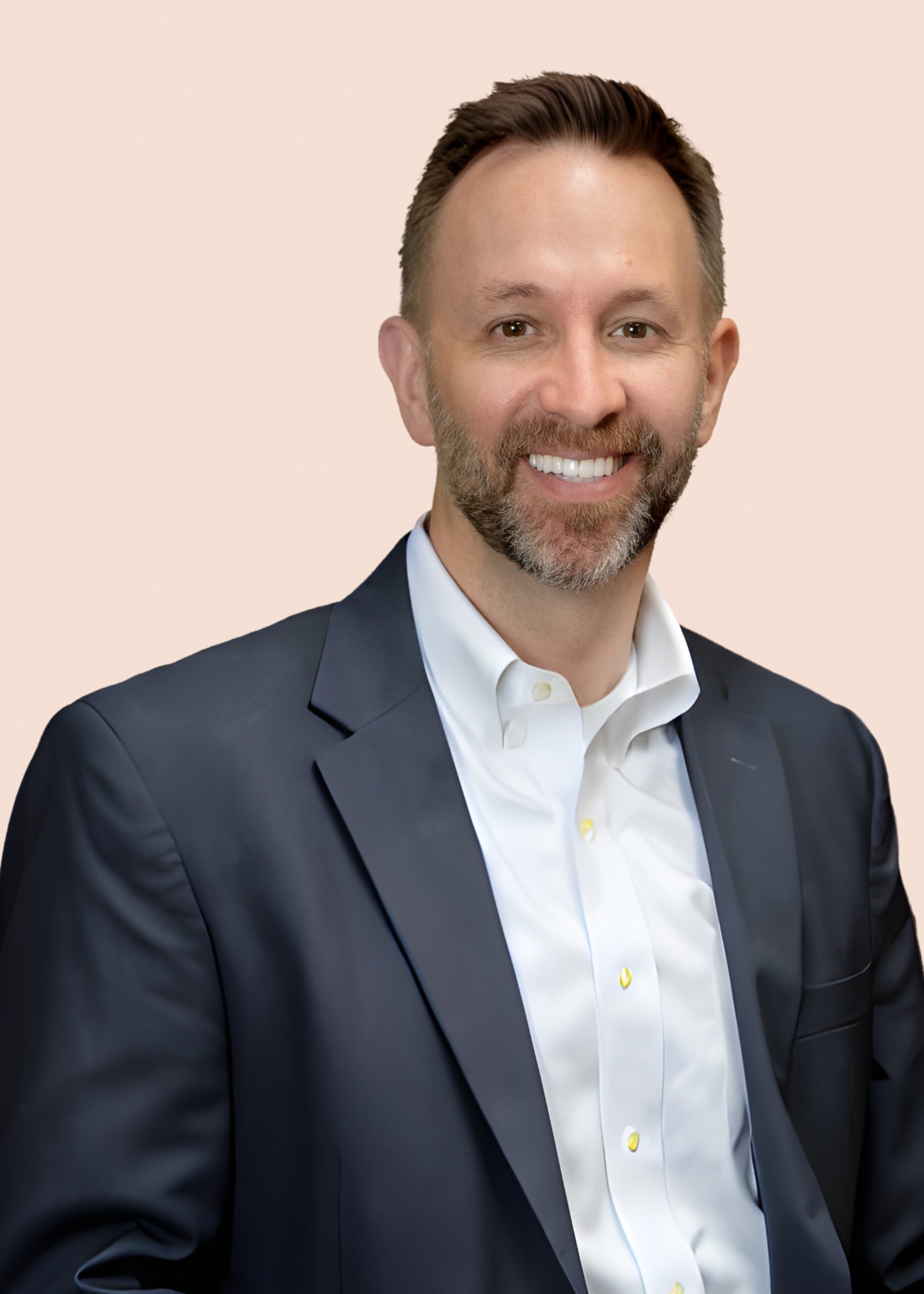 Dr. Schlensker with short brown hair and a beard, wearing a dark suit jacket over a white dress shirt, smiles at the camera against a light beige background.