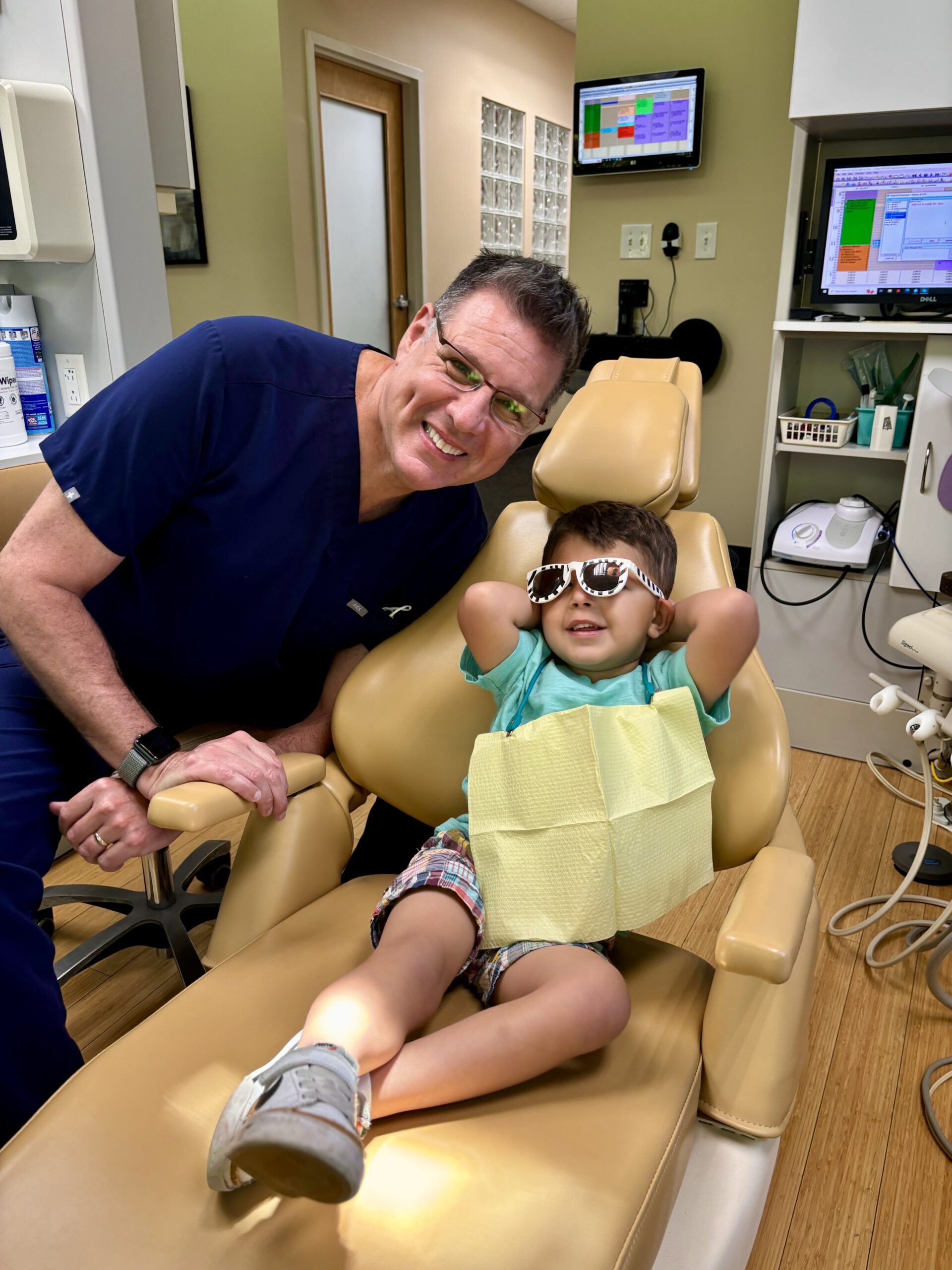 Dr Jose Gallegos in scrubs poses next to a young boy relaxing in a dental chair. The boy wears sunglasses, a dental bib, and casual clothes, looking cheerful and comfortable in the brightly lit dental office.