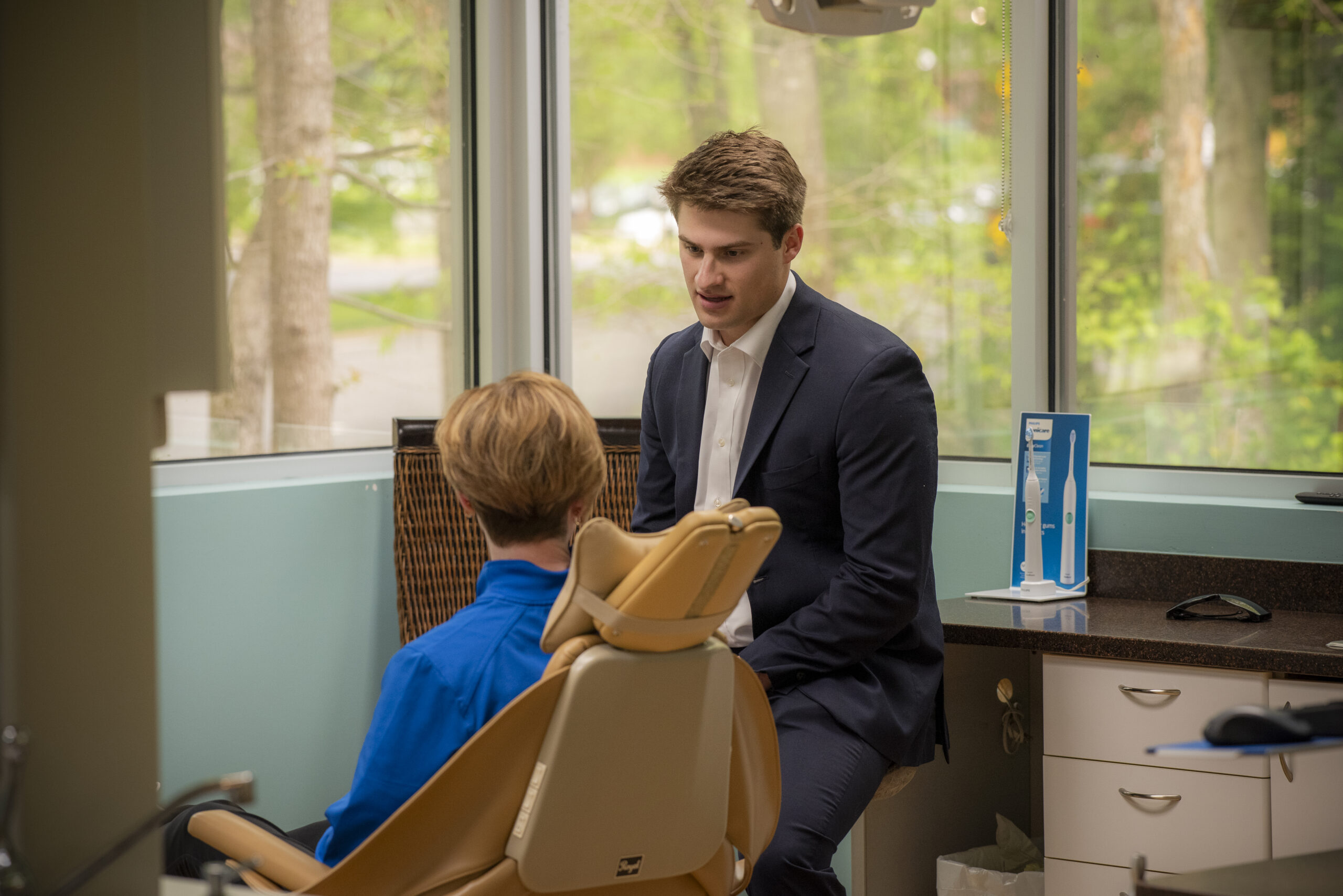 A dentist in a suit talks to a patient sitting in a dental chair by a window in a bright dental office with trees visible outside.