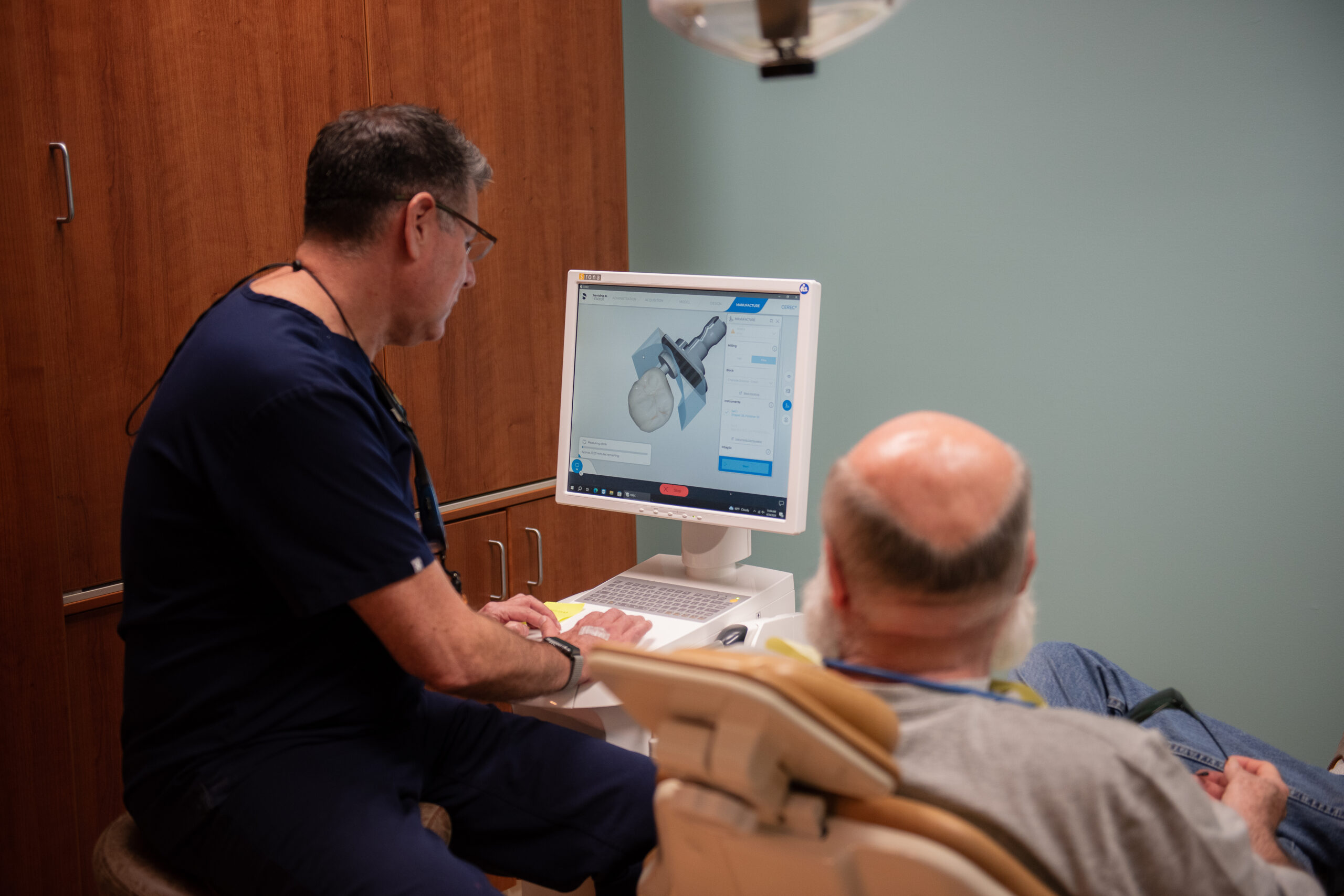Dr Jose in navy scrubs shows a dental scan on a computer monitor to a seated older male patient in a dental office. The computer screen displays a 3D model of a tooth or dental structure.