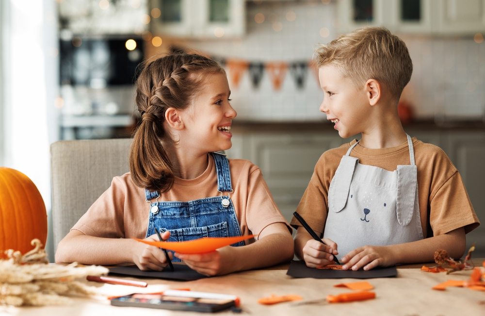 Two smiling children sit at a table crafting with orange paper. One wears denim overalls and holds scissors, while the other, in a light apron, draws on black paper. Autumn decorations and a pumpkin are visible in the background.